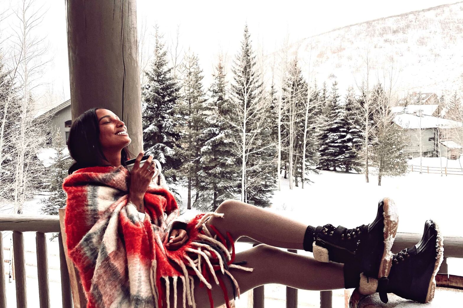 model, jasmine tookes, sits on porch outside wrapped in a blanket and holding a coffee mug with snow covered foliage in the background