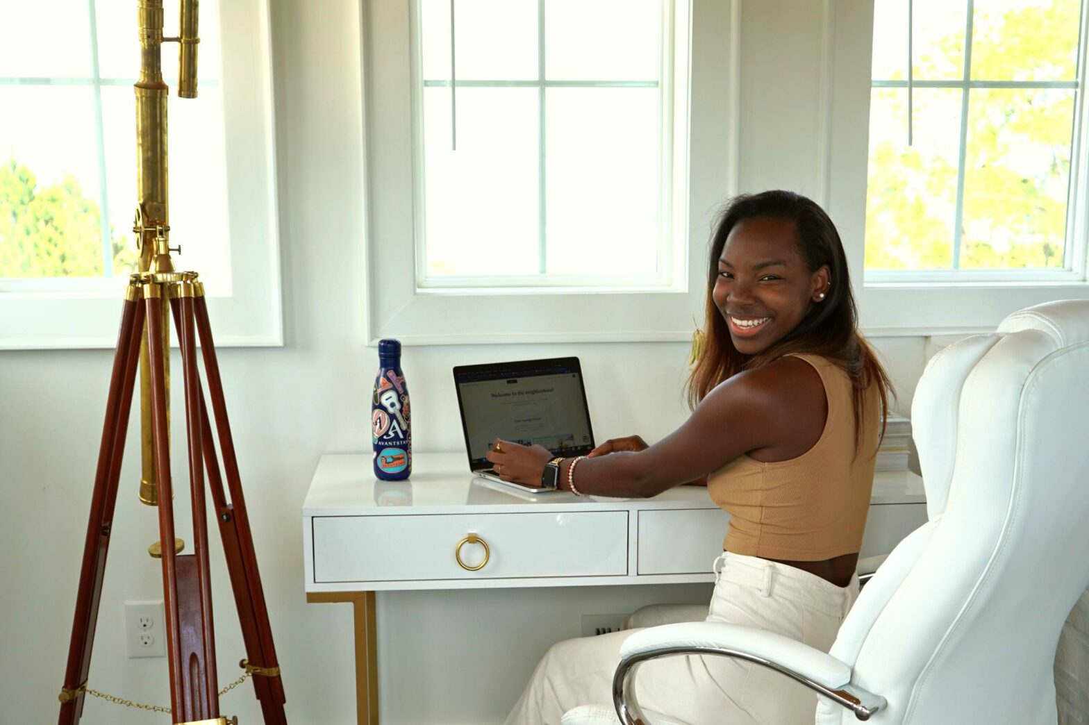 a woman sits at a desk with a laptop open as she looks towards the camera and smiles