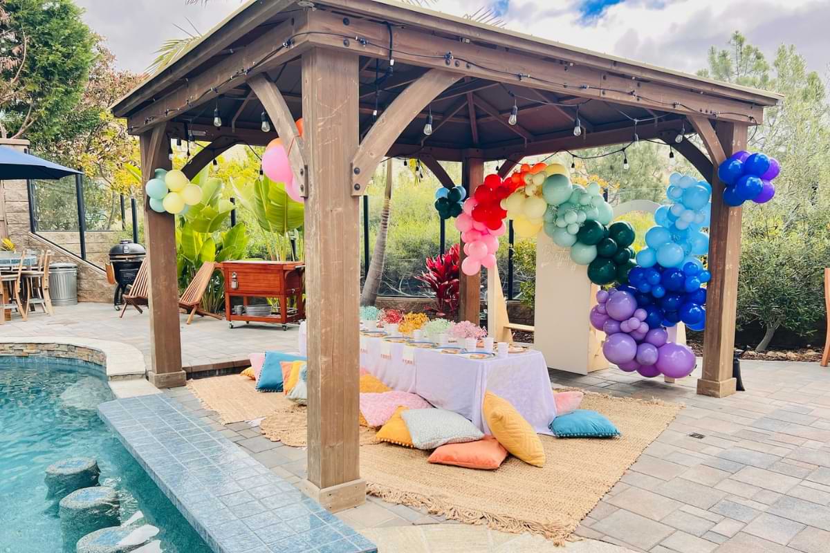 An outdoor knee-height picnic table with birthday decor is set-up with multi-colored pillows on the ground is surrounded by multi-colored balloons under a veranda, next to a pool.
