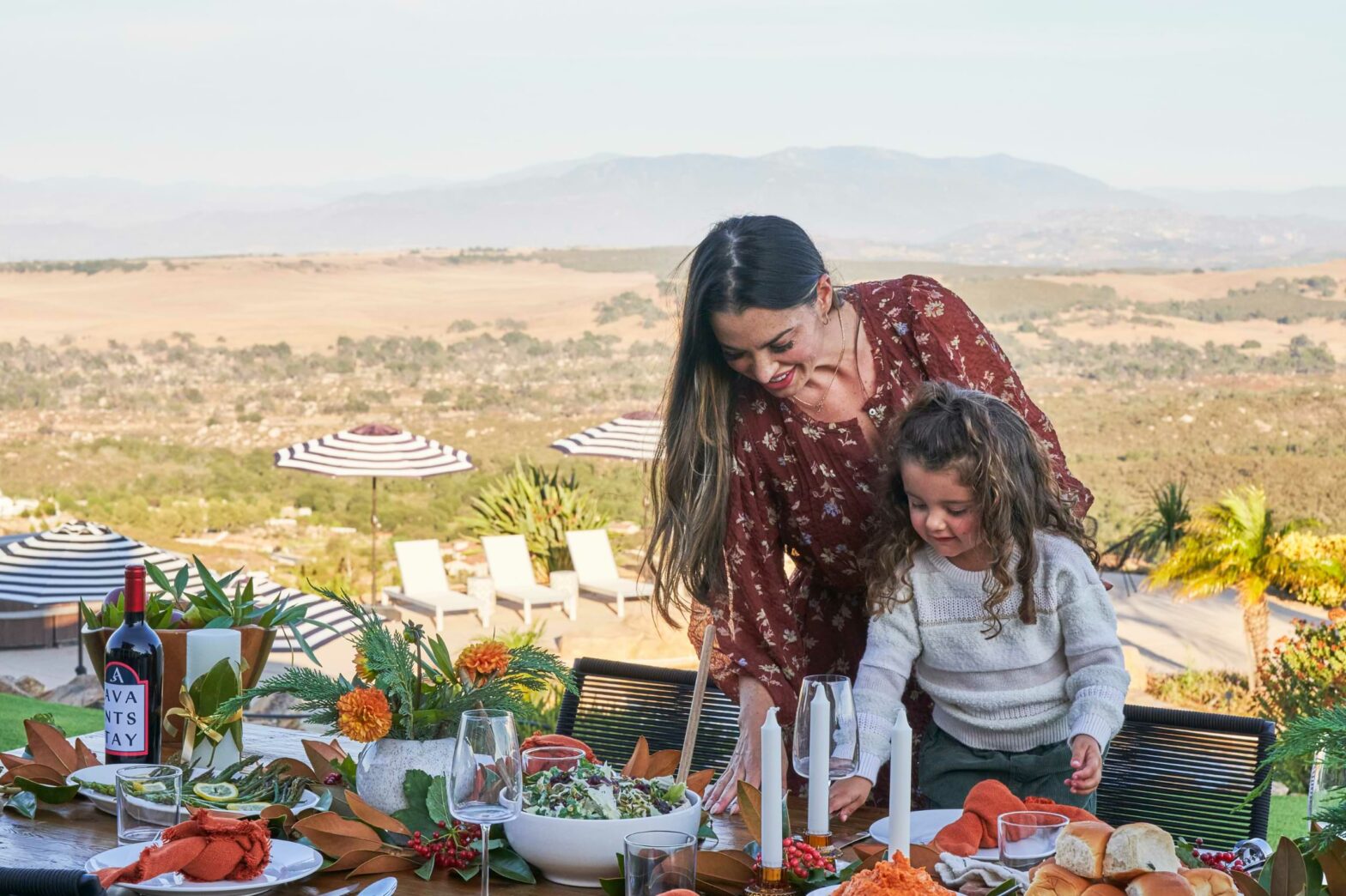 A mother a daughter set a decorated table with fall decor and utensils. In the background, pool chairs and sun umbrellas sit atop a mountain with desert and mountains in the far distance.