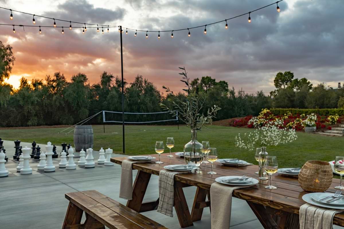 A picnic table with table settings and glasses of white wine sits next to a giant chess set. A volleyball net sits in the background on the open grass while the sun sets in the distance.