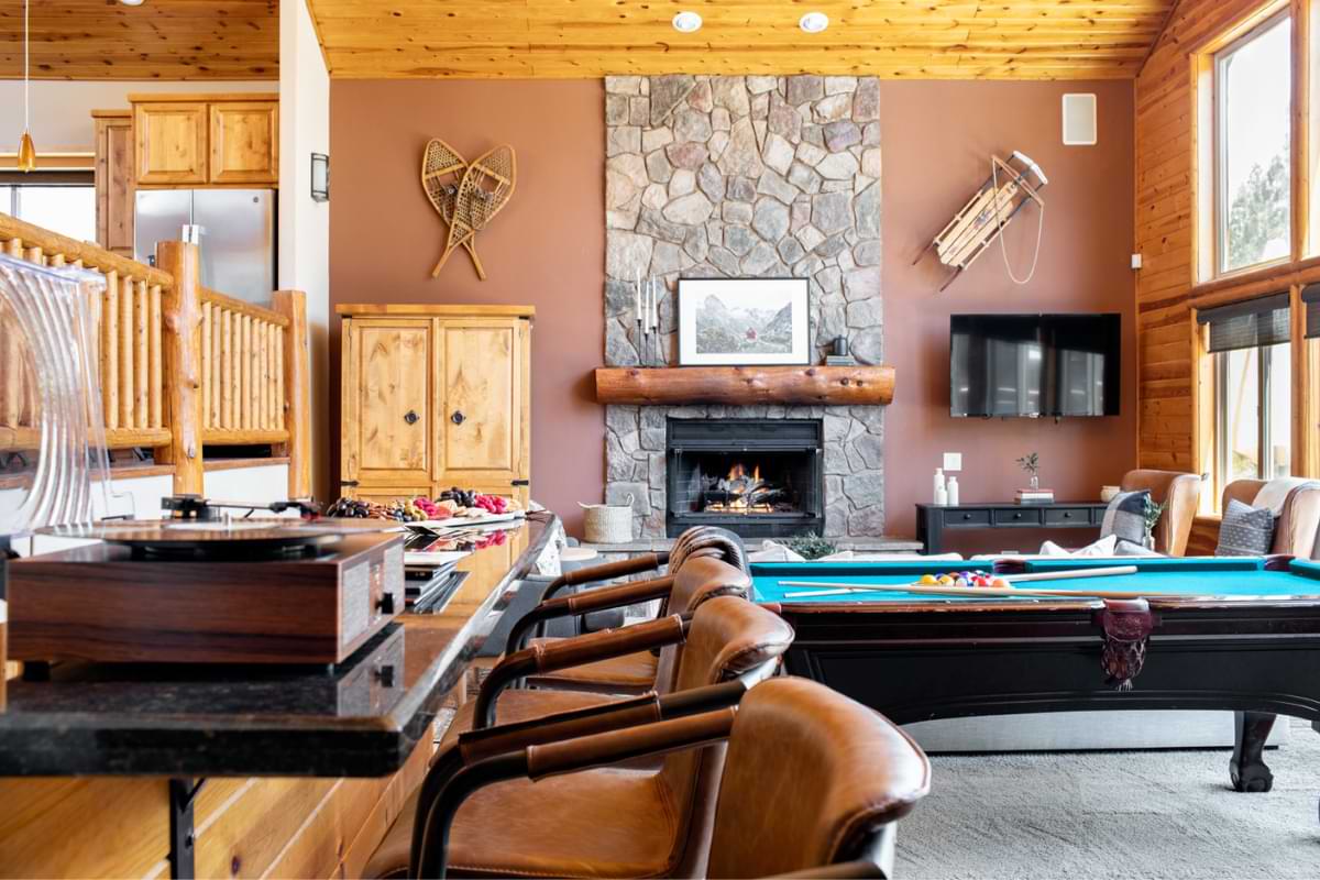 A living room with a pool table, TV, lit fireplace, and wet bar topped with a charcuterie platter and record player. The wet bar has 4 bar-heigh chairs set.