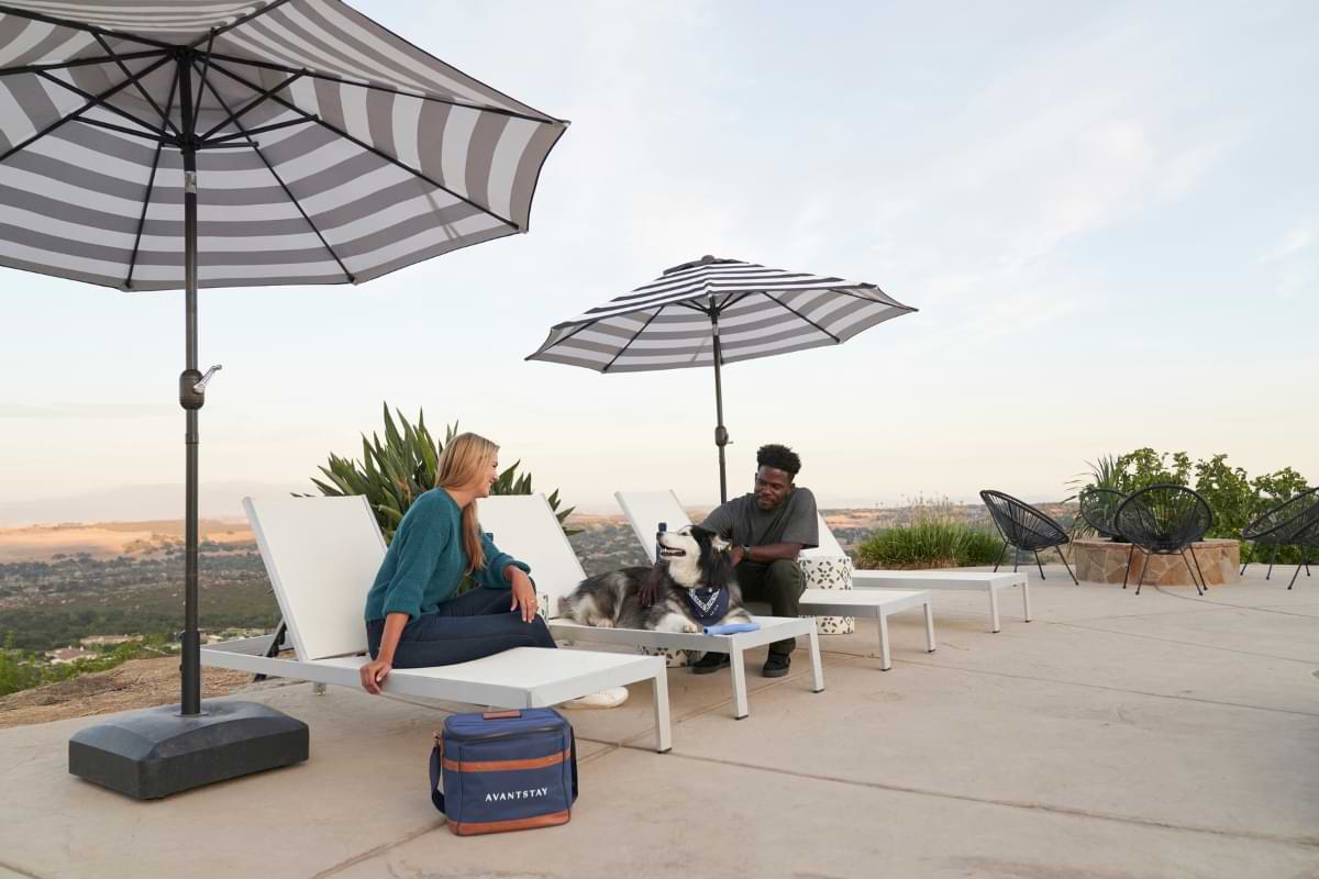 A couple sits in outdoor lounge chairs under sun umbrellas with their large, fluffy dog sitting on a chair in-between them. A cooler bag with "AvantStay" displayed on it sits on the ground.