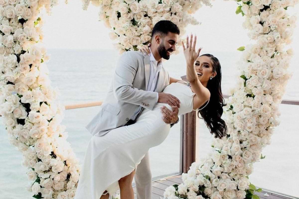 A newly engaged male-female couple in front of a giant heart-shaped arch made of white roses. The woman is being dipped by the man while showing off her engagement ring hand.