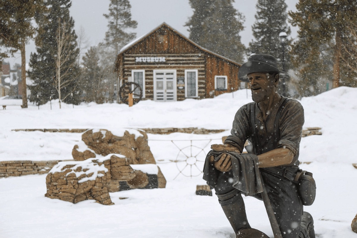A statue of a man sits in the snow outside of a wooden cabin museum in Breckenridge