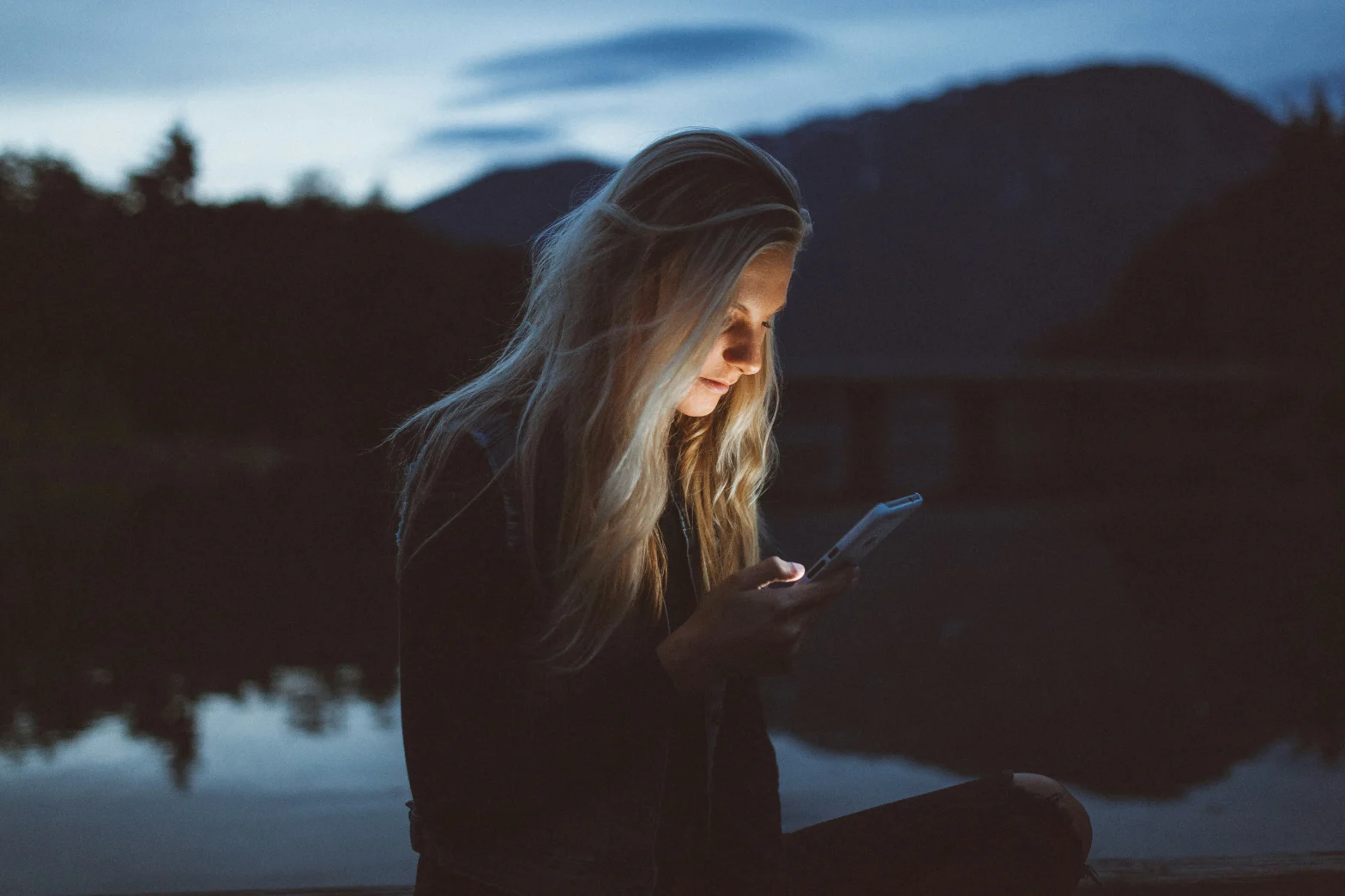 blonde haired girl by the lake by dust checking her phone